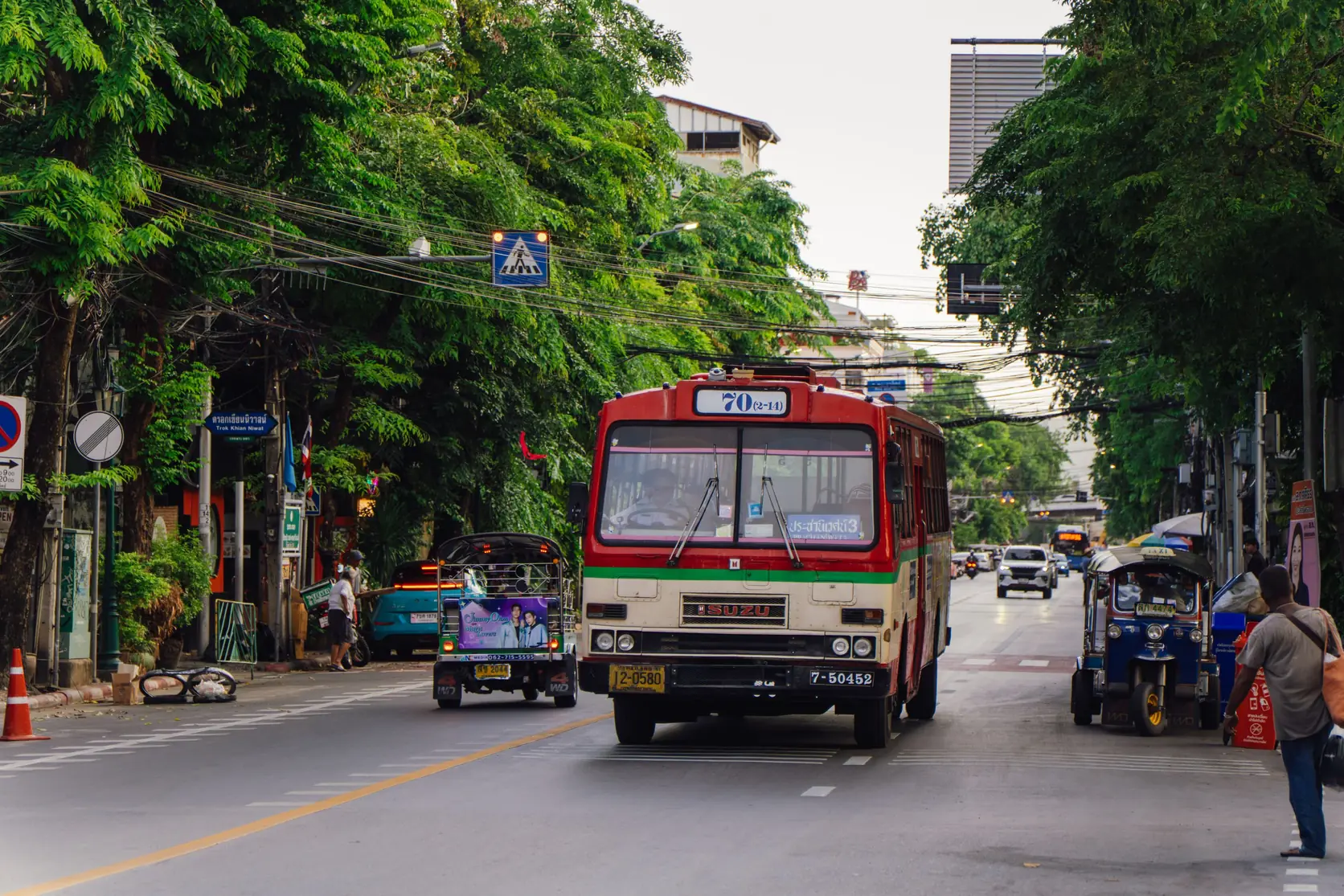 Ein roter Stadtbus und ein Tuk-Tuk auf einer baumgesäumten Straße in Bangkok